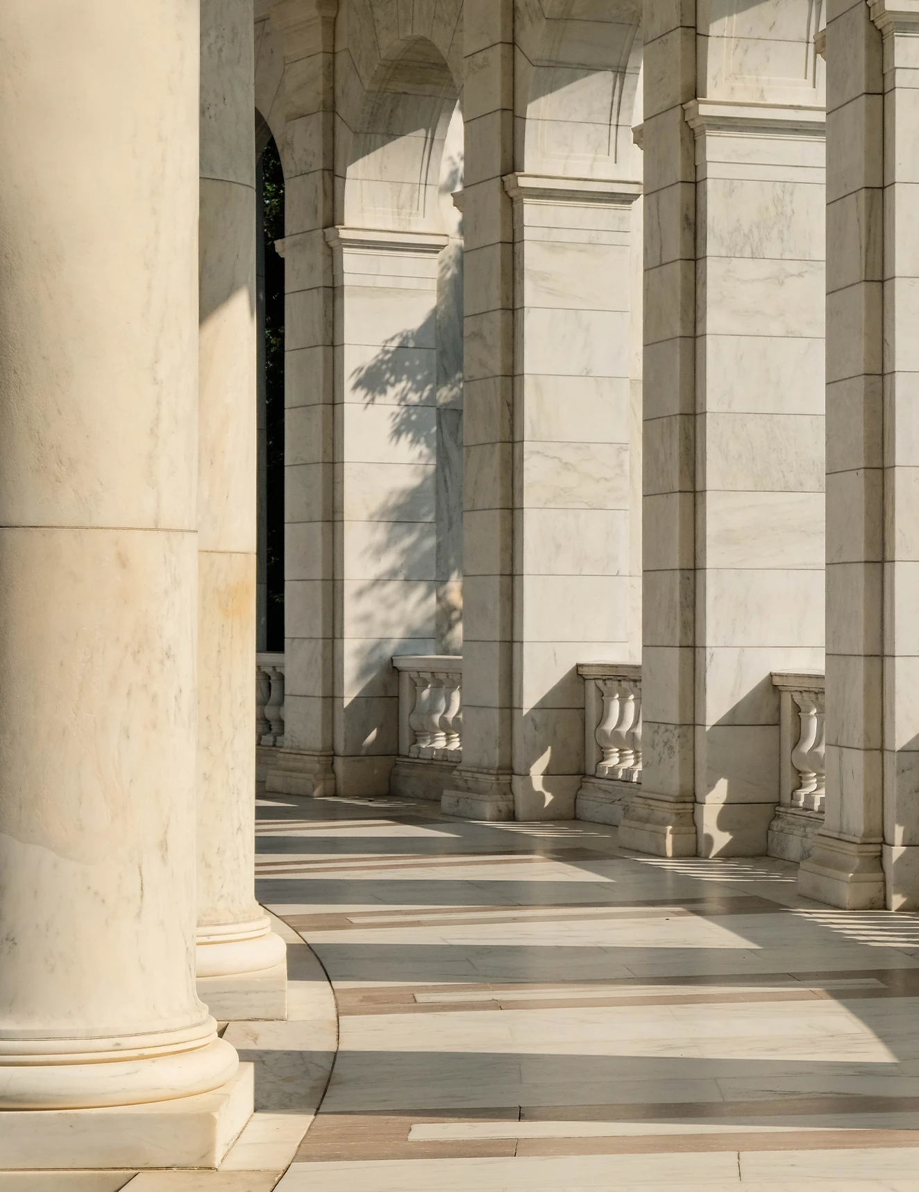 Daylight streaming through a curved collonade. Shadows are cast across the stone path, columns, and archways.