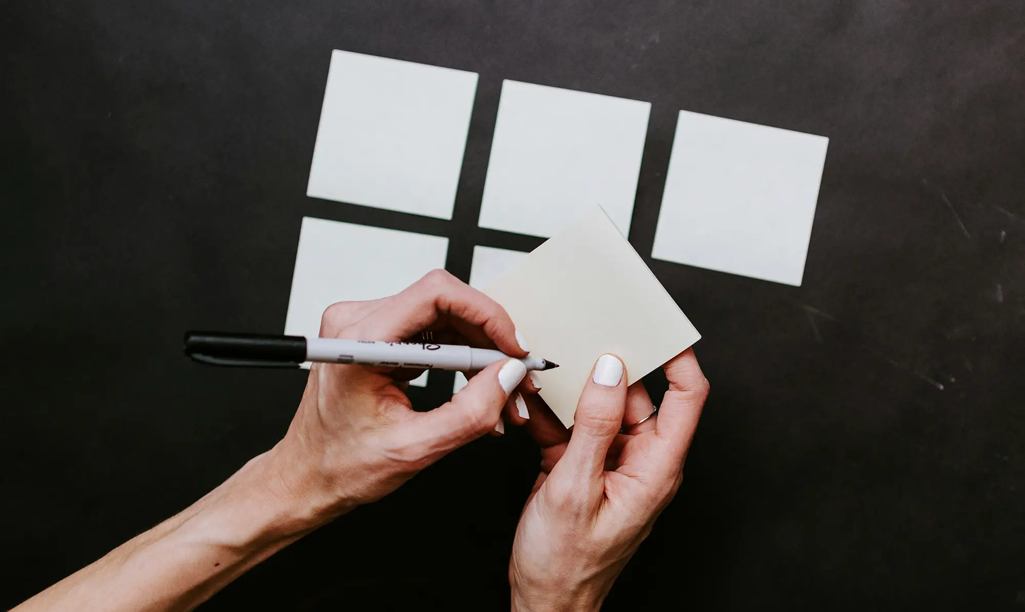 Close up photograph of a woman's hands holding a pen and a blank sticky note. A grid of blank sticky notes are visible in the background.