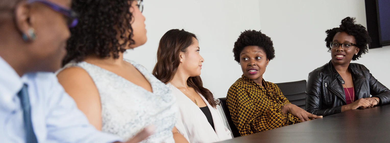 Photograph of a group of people in a meeting. A woman in the center of the image is leading the discussion.
